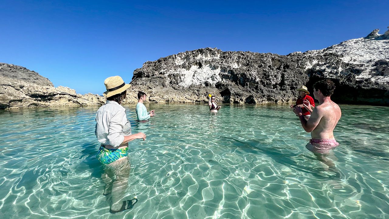 People wading in a clear natural pool beneath limestone cliffs and a blue sky